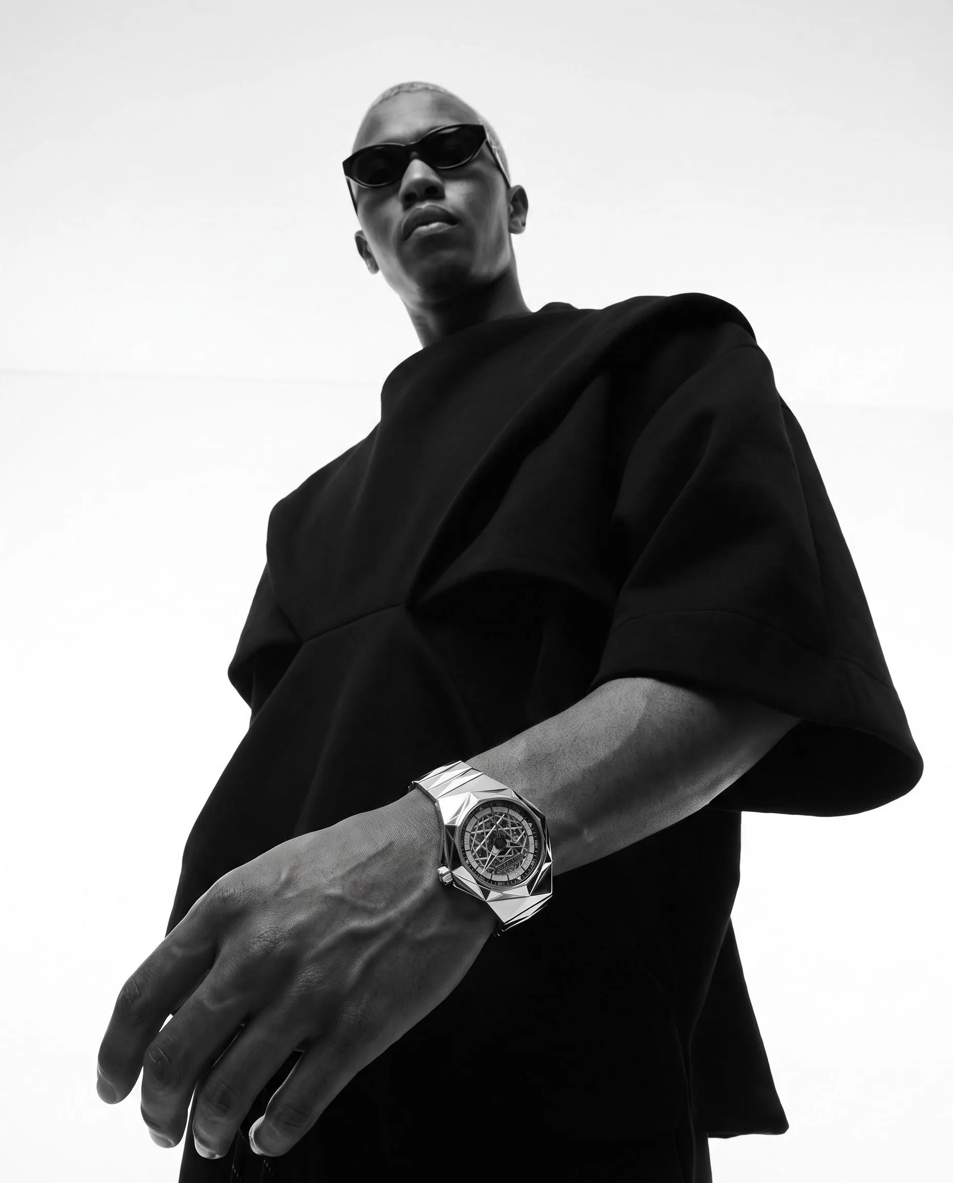 A person wearing dark sunglasses and a black outfit, with a silver bracelet and a large wristwatch, posing against a plain background.