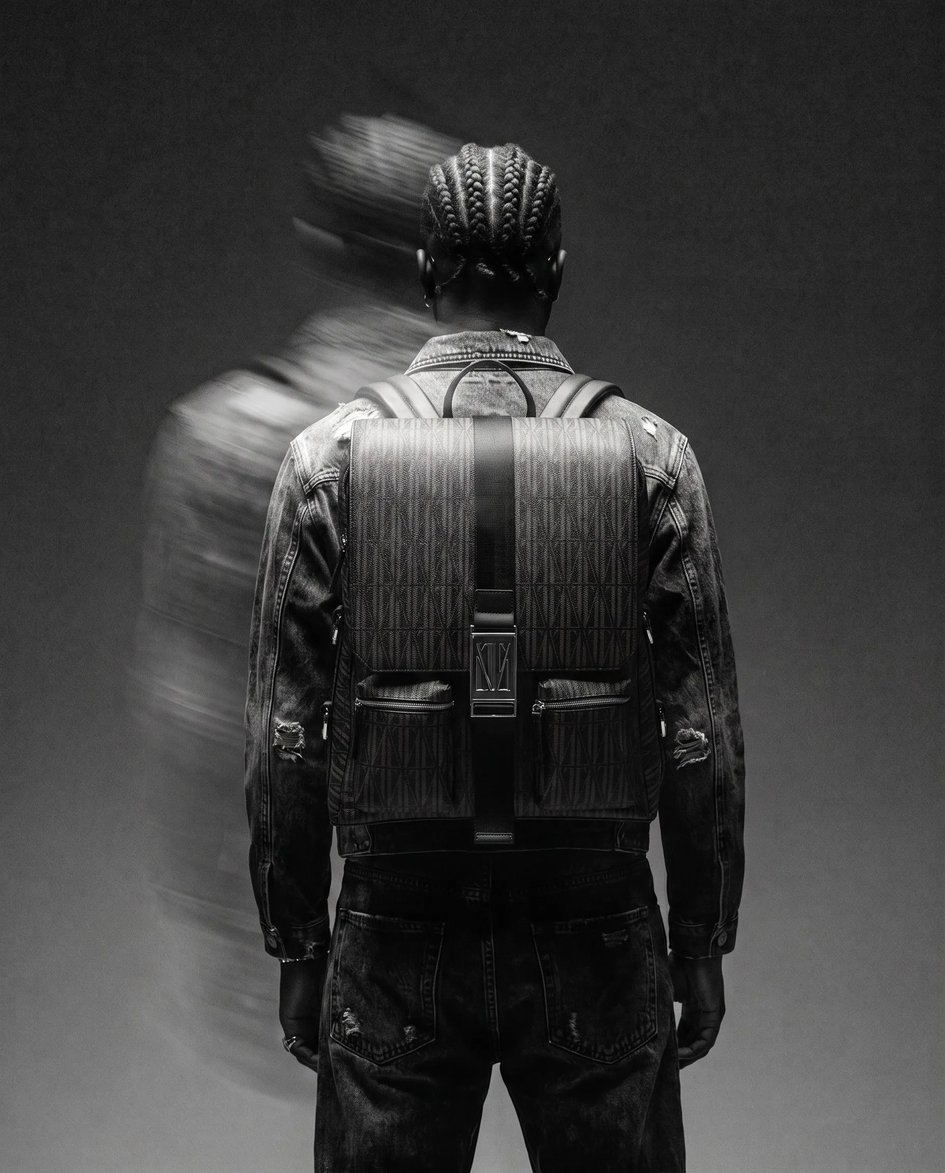 Back view of a person with braided hair, wearing a denim jacket and a large backpack, standing against a plain dark background with a blurred motion effect.