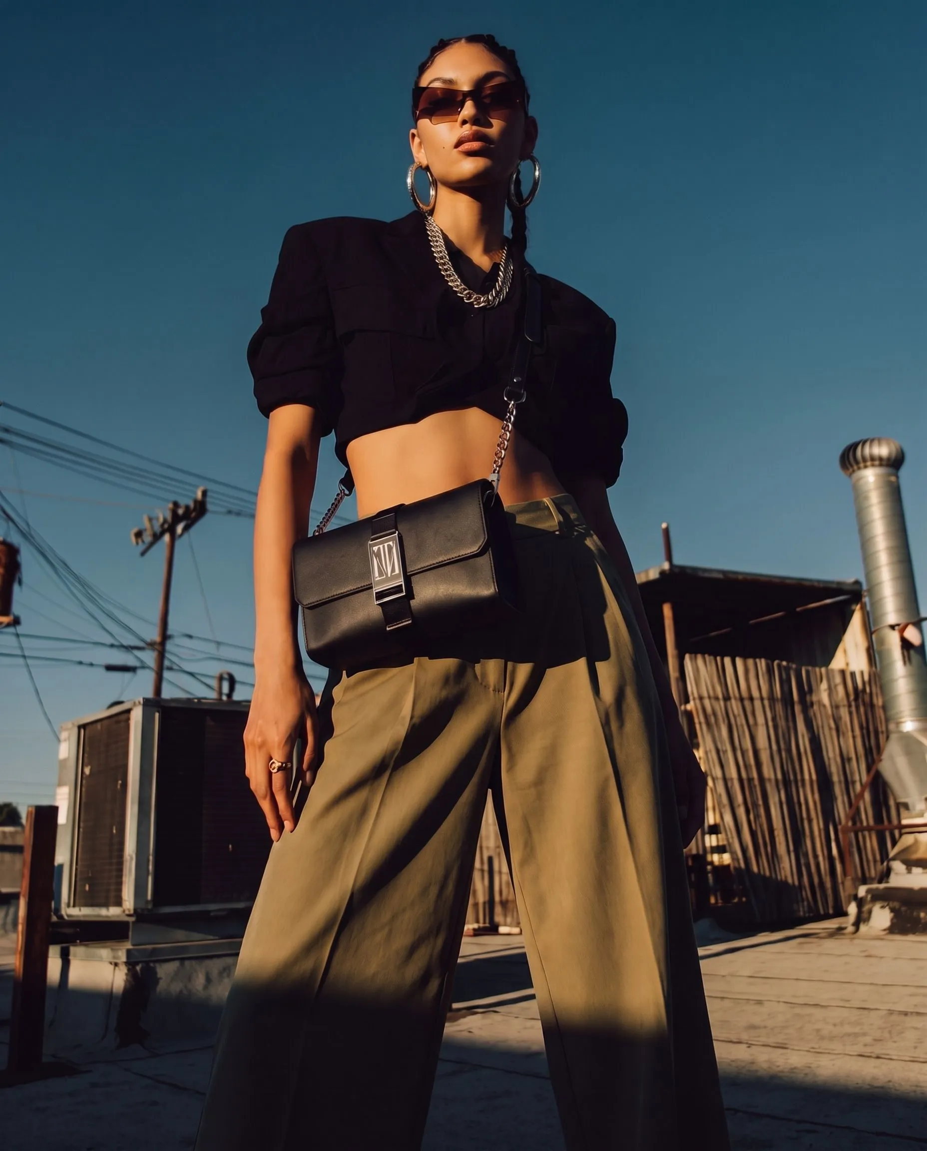 A woman wearing sunglasses, hoop earrings, a chunky chain necklace, a black cropped blazer, and high-waisted khaki pants stands outdoors against a clear blue sky, with power lines and rooftops visible in the background.