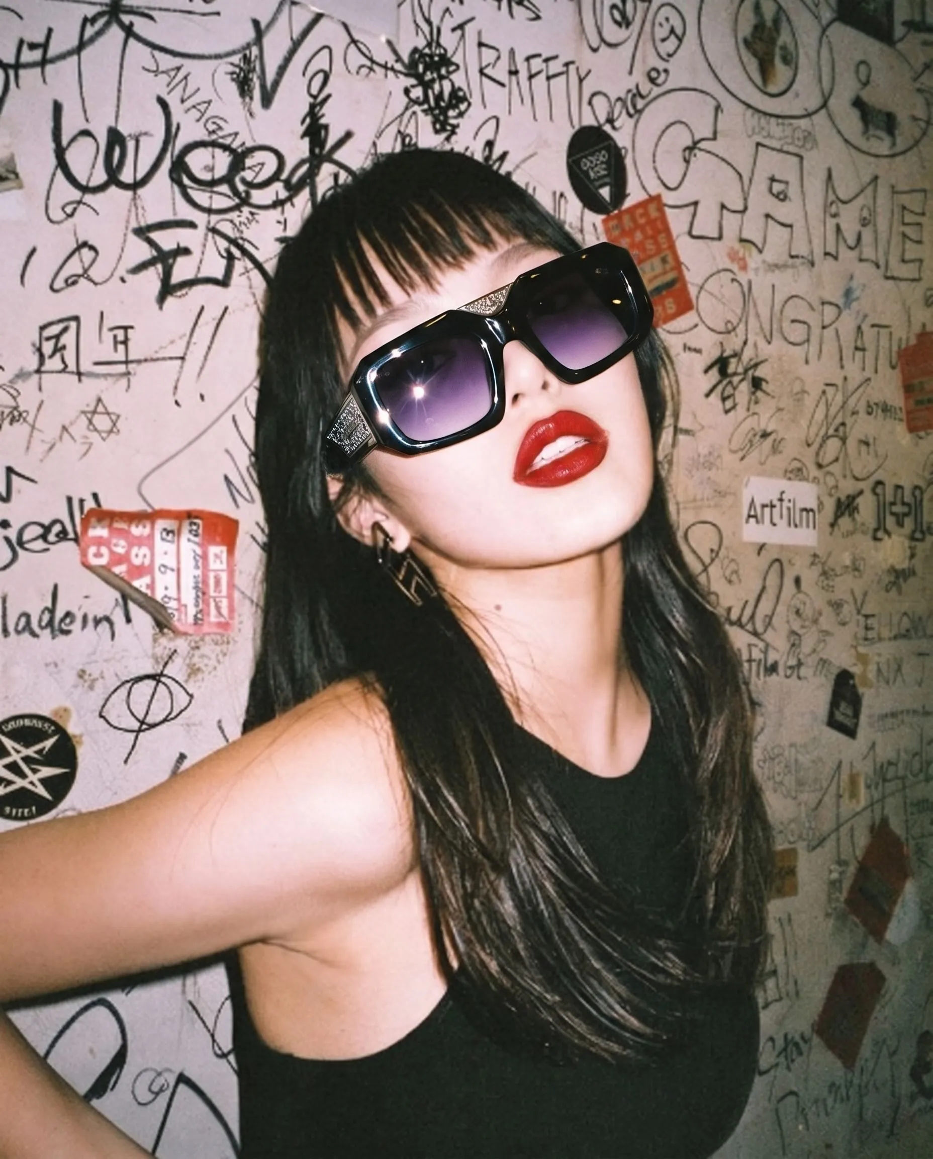 A woman with long dark hair wearing large black sunglasses and red lipstick, standing in front of a wall covered in black graffiti, doodles, and various drawings.