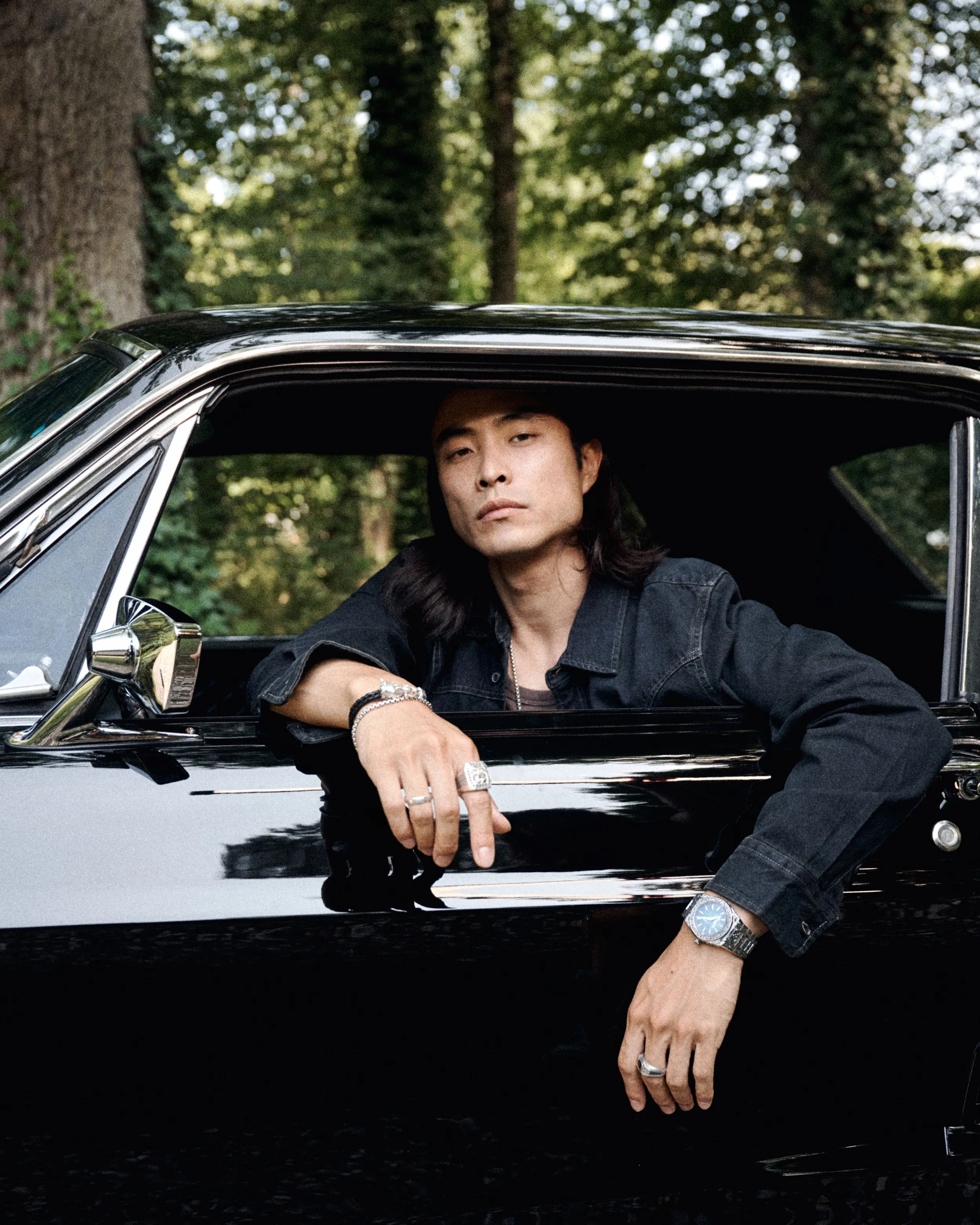 A man with long dark hair leaning out of the window of a black vintage car, wearing multiple rings on his fingers and a silver wristwatch, in a wooded outdoor setting.